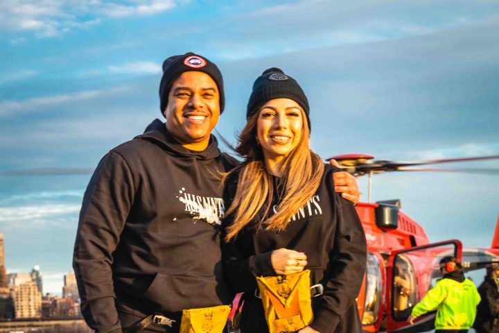 Two people in black outfits pose in front of a red helicopter on a tarmac.