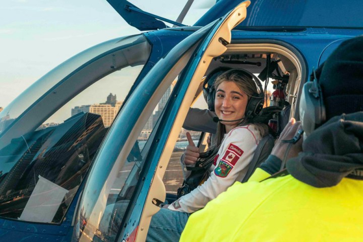 Person in headset sitting in a helicopter cockpit with a thumbs-up, guide in yellow jacket outside.