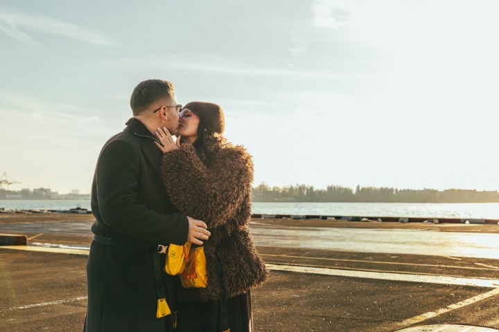 Couple in coats kissing by a waterfront with a sunny sky.