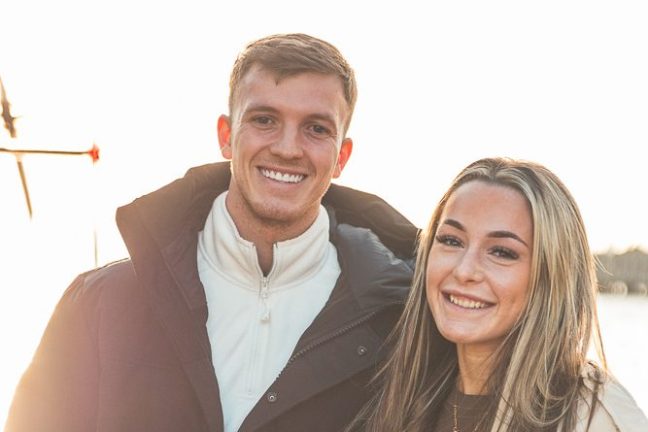 Smiling couple in winter coats outdoors with a bright sky.