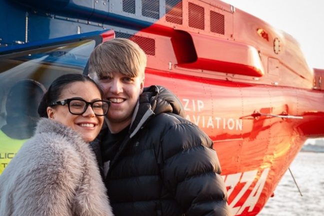 Smiling couple in coats posing in front of a red helicopter.
