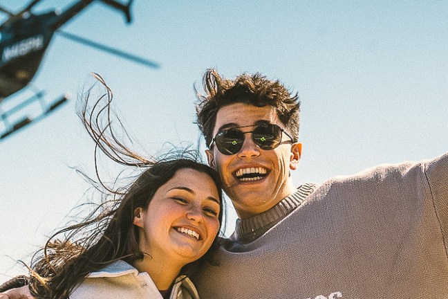 Smiling couple outdoors with helicopter in sky and snack bags, sunny day.