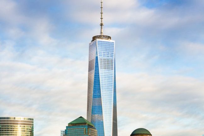 Tall skyscraper with spire in cityscape against cloudy sky and waterfront.