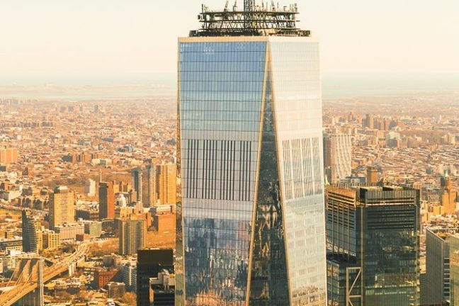 Tall skyscraper with a spire, cityscape in the background under a clear sky.