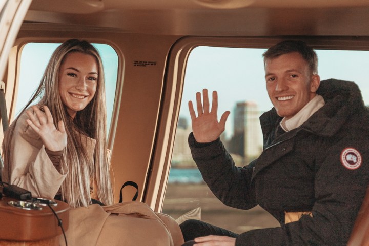 Two people sitting in a helicopter, smiling and waving towards the camera.