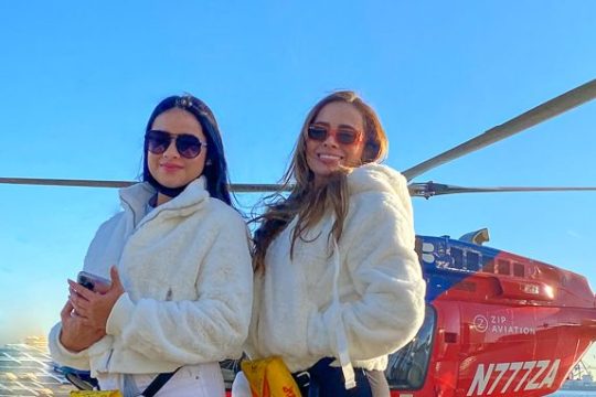 Two women in white jackets and boots posing in front of a red helicopter on a sunny day.