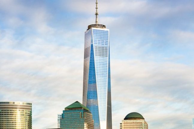 Tall skyscraper with antenna surrounded by buildings near water under cloudy sky.