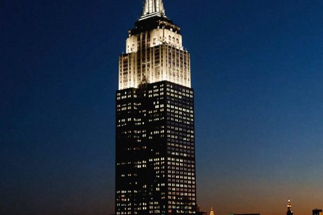 Illuminated skyscraper at night against dark blue sky and cityscape.