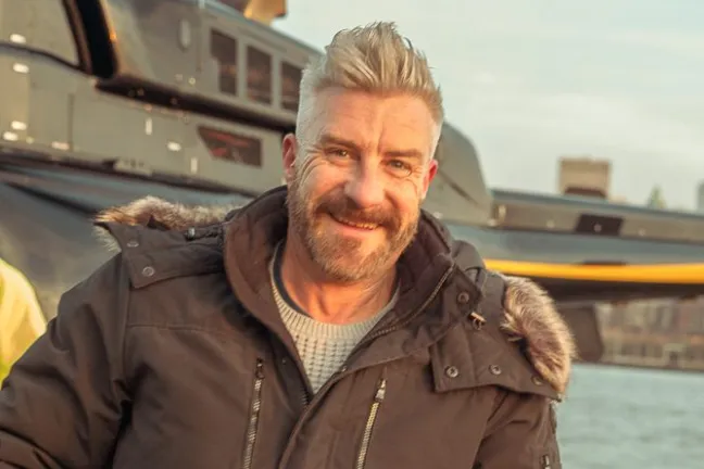 Smiling man in a dark jacket holding a yellow bag in front of a helicopter.