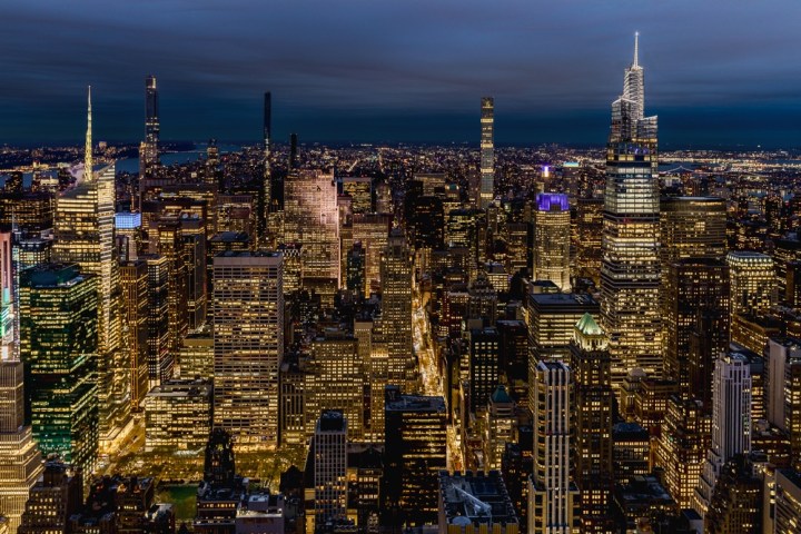 Aerial view of a brightly lit New York city skyline at night with skyscrapers and a deep blue sky.