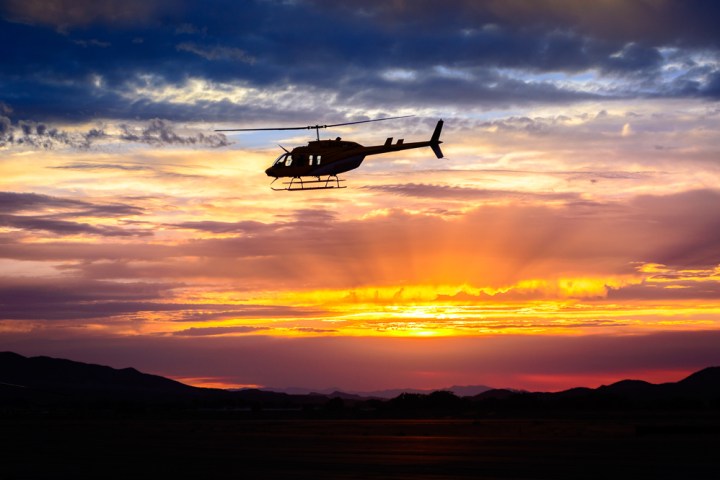 Silhouette of helicopter flying at sunset with colorful sky and mountains in background.
