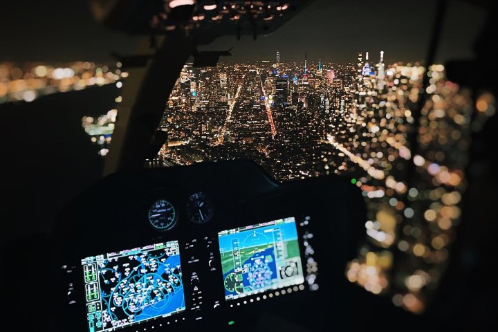 Helicopter cockpit view over a city skyline at night, with illuminated buildings and navigation screens visible.