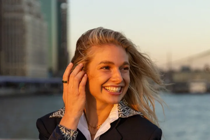 Smiling person with long hair and a blazer, standing on a helipad by a river at sunset.